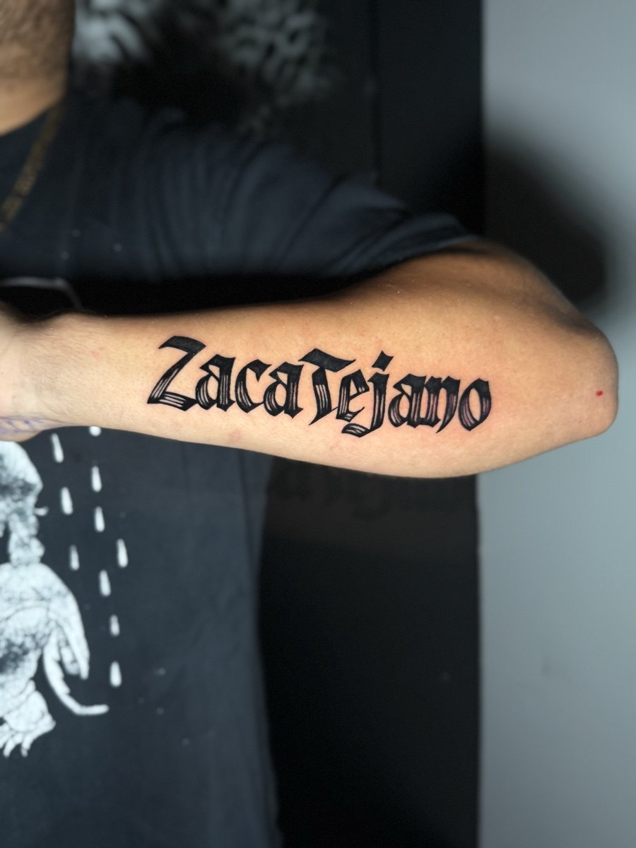 Bold blackletter name tattoo along a forearm.